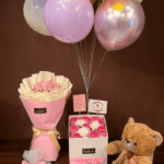 Luxury gift set featuring a brown teddy bear, chocolate bouquet, birthday roses in a box, and colorful balloons against a dark brown background.