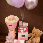 Luxury gift set featuring a brown teddy bear, chocolate bouquet, birthday roses in a box, and colorful balloons against a dark brown background.